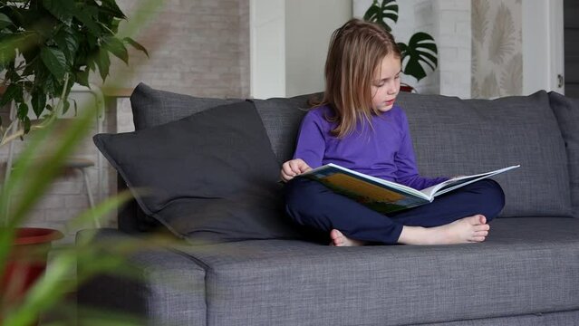 a little blonde girl is reading a big book sitting on the sofa in the living room.Smile and interest in reading.Self-education of children, reading literature at home, interesting leisure.
