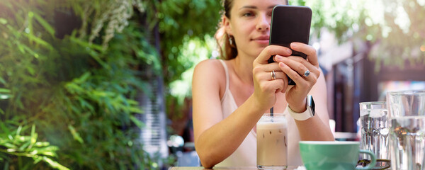 Smart phone in hands of young woman sitting in sidewalk cafe in summer on background. 