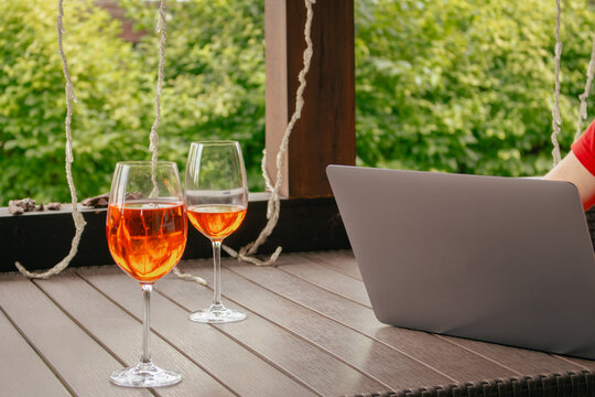 Laptop with glass of Aperol cocktail. Unknown person working on wireless computer in outdoor cafe. Freelancer in public place. Grey laptop and two glasses of wine on table. Tranquil vacations. 