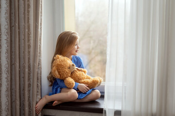 A little girl with long blond hair sits on the windowsill with a toy and looks out the window into the street. This is a sad mood.