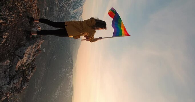 Pride month LGBTQ. Young oman waving LGBT gender identity flag at the mountain peak at sunset. Vertical shot