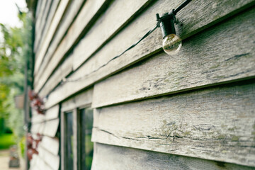 Shallow focus of one of many hanging festoon lights seen hanging around a rustic wooden barn. The lights are party of a wedding celebration at a private garden party.