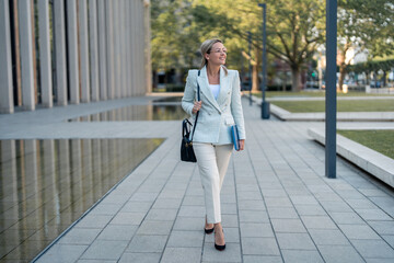 Smiling confident businesswoman with laptop walking outside the building and looking away.