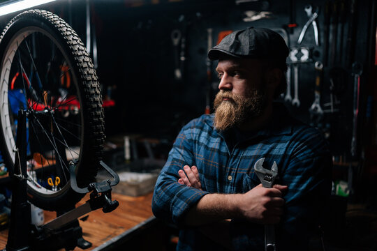 Portrait of thoughtful bearded repairman in cap holding wrench standing with crossed arms by bicycle in repair workshop with dark interior, looking away. Concept of professional bicycle maintenance.