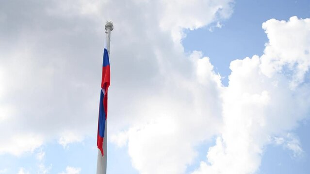 Low angle view of Russian national flag hanging on large flagpole against blue sky with white clouds in sunny summer windless day. Real time handheld video. Copy space. Politic symbols theme.
