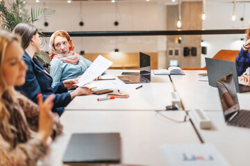 Young woman in hijab listening to her brunette female colleague explaining profit report at work
