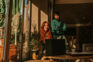 Handsome male person standing behind his female ginger colleague and watching her working on the lap top. He is giving suggestions