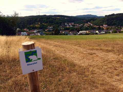 Schild vom Premium-Wanderweg Biberpfad mit Blick auf den Ort Berschweiler in der Gemeinde Marpingen im Landkreis St. Wendel, Saarland.
