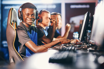 Side view of a cheerful african guy, professional cybersport gamer wearing headphones looking at camera and smiling while participating with team in eSport tournament © Friends Stock
