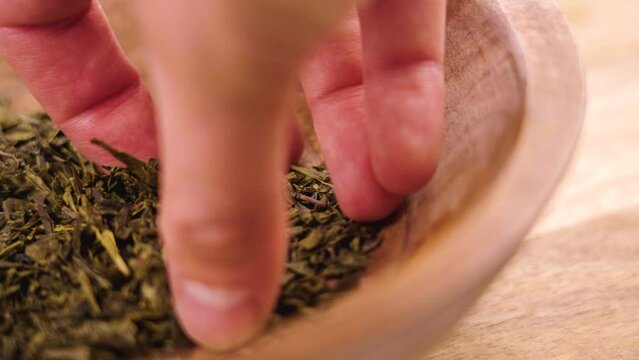 Hand taking a handful of dried green japanese sencha tea leaves from a rustic bowl in slow motion closeup
