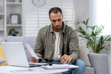 Latin American man is focused working remotely at home on a laptop and with documents