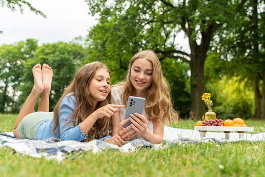 Two cute blonde girls lie in the park on a blanket on the grass, holding the phone in hands, browsing the news feed. Girls on a picnic chatting, touching the phone screen, scrolling and swiping