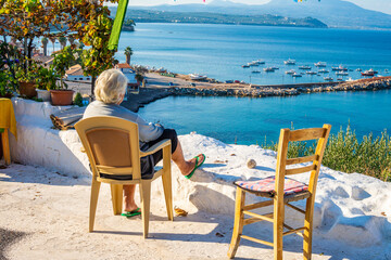 Old woman sitting on a chair gazing the amazing view of Koroni coastal village. Beautiful scenic in Koroni, Greece, Europe.