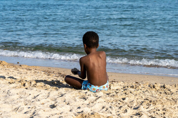 A dark-skinned boy sits with his back on the seashore near the water and plays with sand.