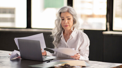 Upset Businesswoman Looking Through Papers Sitting At Workplace Near Laptop