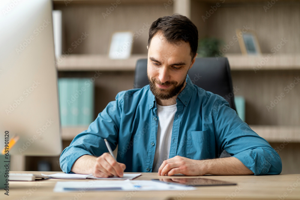 Poster smiling handsome businessman taking notes to notepad at workplace in white office
