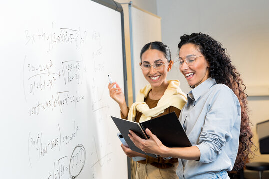 Two beautiful female students of Eastern appearance stand in a classroom near the blackboard solve mathematical equations look at the Notebook smile see the correct solution. Teamwork leads to success