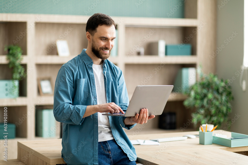 Poster Smiling Businessman Using Laptop Computer While Standing Near Desk In Modern Office