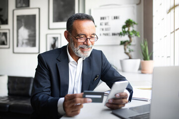 Cheerful caucasian senior businessman in suit, glasses pay at phone at table with computer, uses credit card