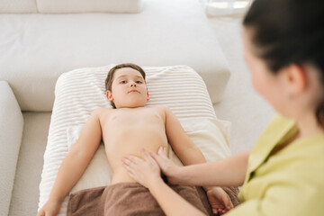 Obraz na płótnie Canvas High-angle view of five year old boy patient having therapeutic massage on tummy lying on couch in medical clinic. Closeup hands of unrecognizable female pediatric masseuse massaging tummy of child.