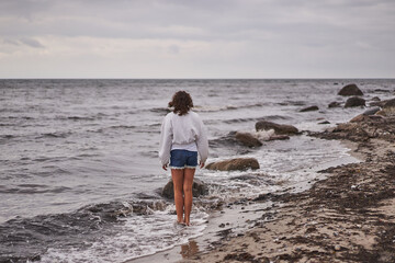 Fototapeta na wymiar Teenage girl walking alone on the sand beach of North sea in Denmark during her summer holidays or vacation and watching horizon. Picture is taken during cloudy and windy summer scandinavian evening.