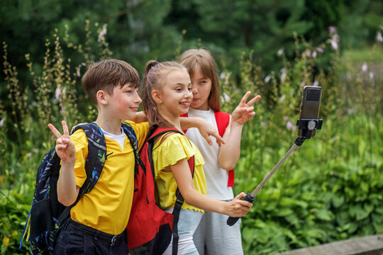Group children with backpacks taking picture by smartphone on selfie stick outdoor. Playtime.