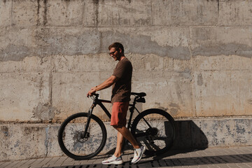 Fototapeta na wymiar Full length of handsome young man pulling his bicycle against a concrete wall outdoors