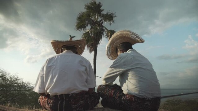 Two people sitting under the palm tree, playing string instrument of sasando in the afternoon