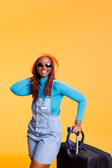 Fototapeta na wymiar Female model posing with sunglasses and hat in studio, carrying suitcase trolley to leave on vacation trip. Smiling woman feeling excited about urban adventure abroad, joyful lifestyle.