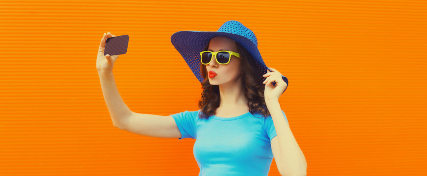 Stylish young woman 20s taking selfie with smartphone and blowing her lips wearing straw hat on orange background