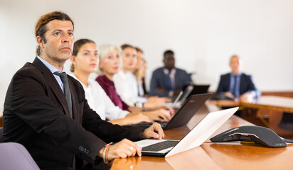 Fototapeta premium Man in suit sitting at table with his colleagues, attending meeting and using laptop.
