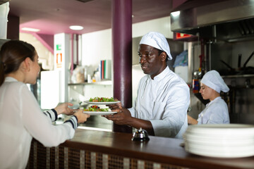 Fototapeta na wymiar Focused young adult man chef giving out ready meals to waitress on order station in open restaurant kitchen