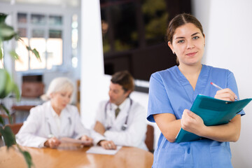 young medical female colleague standing in meeting room and employees are talking behind her at table