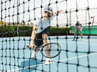 Portrait of young man tennis player during doubles couple match at court. View through the tennis net