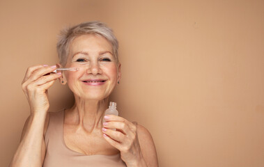 Happy senior lady applies cosmetic oil serum on face takes care of skin and smiles broadly enjoys...