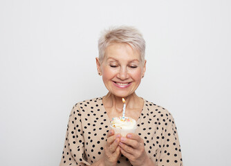 Cheerful elderly woman with short haircut holds a cupcake with a candle in her hands, celebrates a birthday and laughs