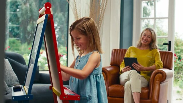 Family with daughter drawing picture on easel as mother with digital tablet watches from sofa at home -shot in slow motion 