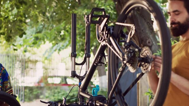 Fit and dedicated multiracial pair inspecting damaged bicycle for maintenance using professional equipment. Boyfriend mending bike pedal and talking with girlfriend who is removing broken wheel.