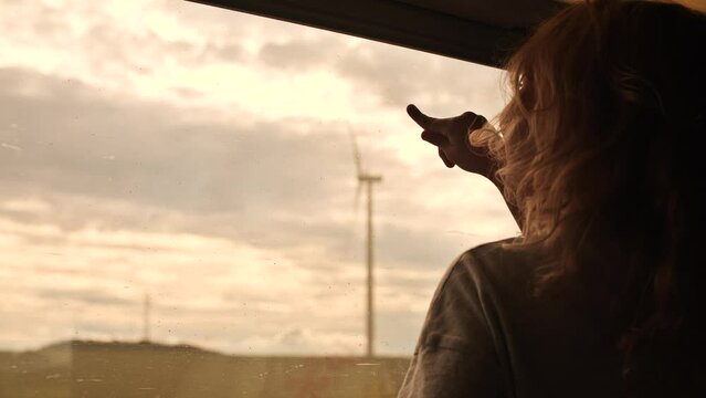 Happy little blonde girl looks through the train window at modern wind turbines