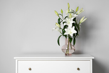Beautiful bouquet of lily flowers in glass vase on chest of drawers near white wall, space for text