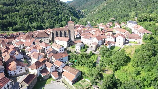Scenic aerial view of spanish township of Isaba with brownish tiled roofs of residential buildings and medieval fortified church of Saint Cyprian in Roncal valley surrounded by green Pyrenees, Navarre