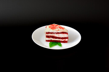 A piece of Red velvet cake with mint leaves on a round and white porcelain plate on a black background.