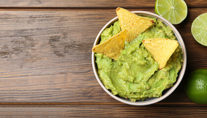 Bowl of delicious guacamole, lime and nachos chips on wooden table, top view. Space for text