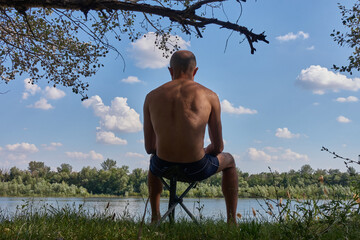 Wet, after swimming, a young man in swimming trunks sits on a folding chair on the river bank and looks at the water. Sky in the clouds.