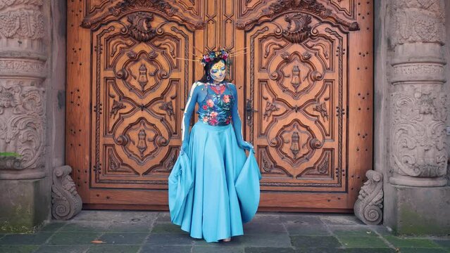 Dancer in costume for dia de los Muertos, woman with long dark hair in traditional costume and Mexico Catrina makeup standing near wooden ornamental door and holding skirt. Day of death.