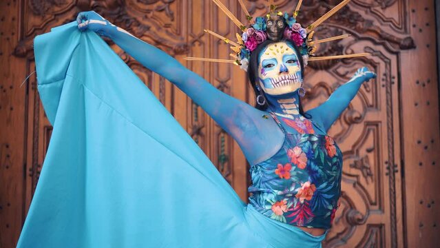 Dancer in costume for dia de los Muertos, woman with long dark hair in traditional costume and Mexico Catrina makeup standing near wooden ornamental door and holding skirt. Day of death.
