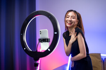 Young trendy influencer asian woman dancing on mobile phone at home in living room with neon light. Creator vlogger talent dancing enjoy hobby content recording show video sharing on social media.