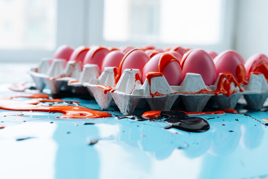 Side view of pink eggs in white carton partly covered with thick orange paint, its flows running down on blue table