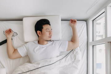 Asian man falling asleep with TV remote control and eyeglasses on white bed.