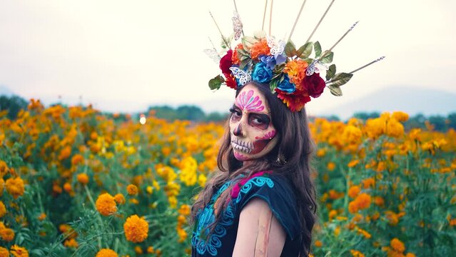 Woman with Mexico Catrina makeup in flower field, serious woman in traditional costume and headwear wearing, standing on field among blooming marigold flowers while looking away. Day of death.